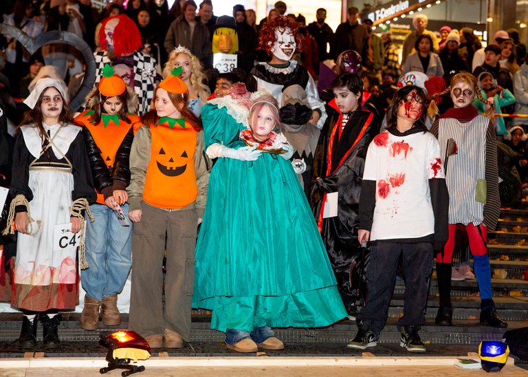 Gruppen poserar i olika Halloween-kostymer på en scen med publik i bakgrunden. Kostymerna inkluderar jack-o'-lantern-motiv, blodfläckar, teaterliknande kläder och skräckmasker.