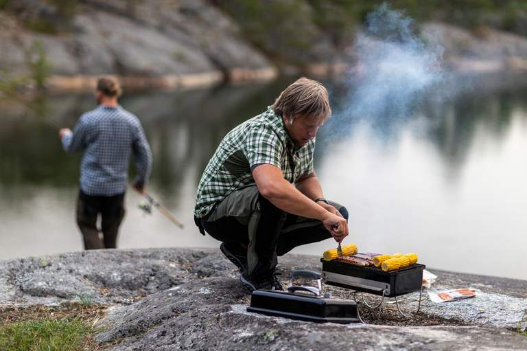 A man is grilling corn on a small portable grill, among the cliffs on the island of Hjälpskär.
