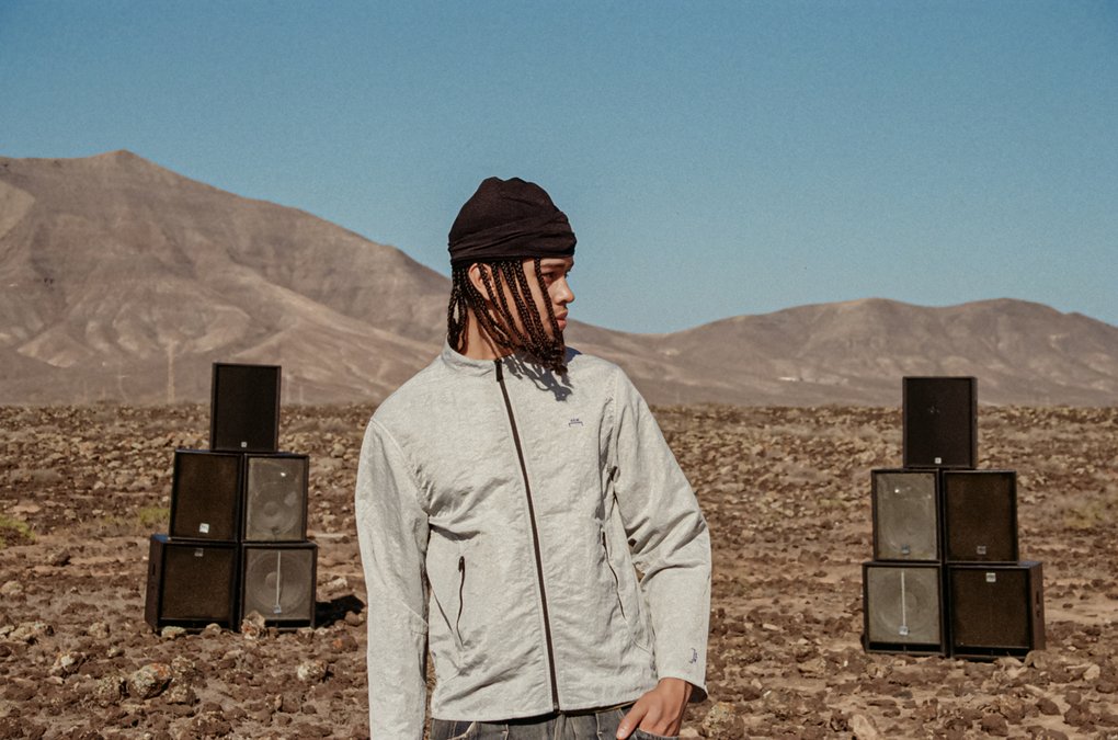 En man i ljusgrå jacka, med en uppsättning högtalare bakom sig, står i ett ökenlandskap med en bergskedja i bakgrunden.