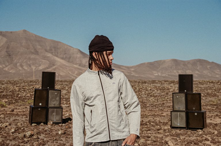 En man i ljusgrå jacka, med en uppsättning högtalare bakom sig, står i ett ökenlandskap med en bergskedja i bakgrunden.