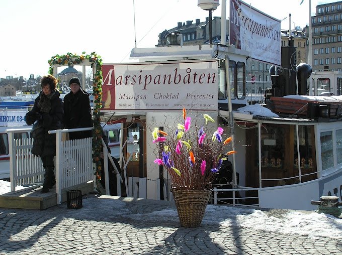 Marsipanbåten förtöjd vid Nybrokajen i Stockholm med två besökare i tjocka jackor som går av landgången. Framför står ett påskris med fjädrar. Framför båten på kajen står ett stort påskris med färgglada fjädrar.