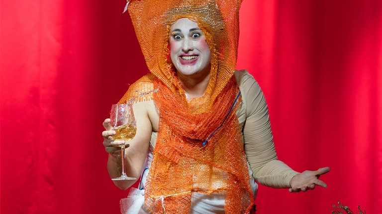 En person klädd i en orange, genomskinlig plastfolie med en liknande hatt håller ett glas med dryck.