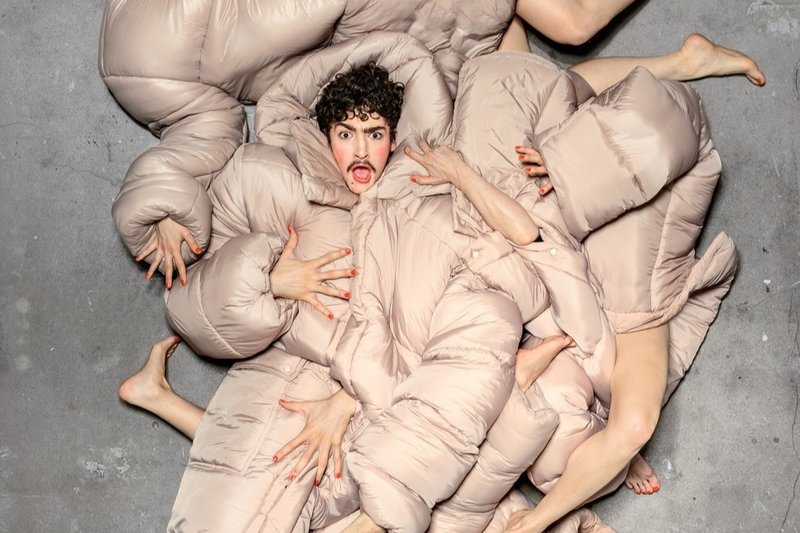 En person ligger på ett betonggolv omgiven av flera kroppar insvepta i beige, vadderade täcken. Armar, ben och händer sticker fram i olika riktningar och skapar en sammanflätad, skulptural formation. Personen i mitten har ett uttrycksfullt ansikte med öppen mun och blicken riktad uppåt.