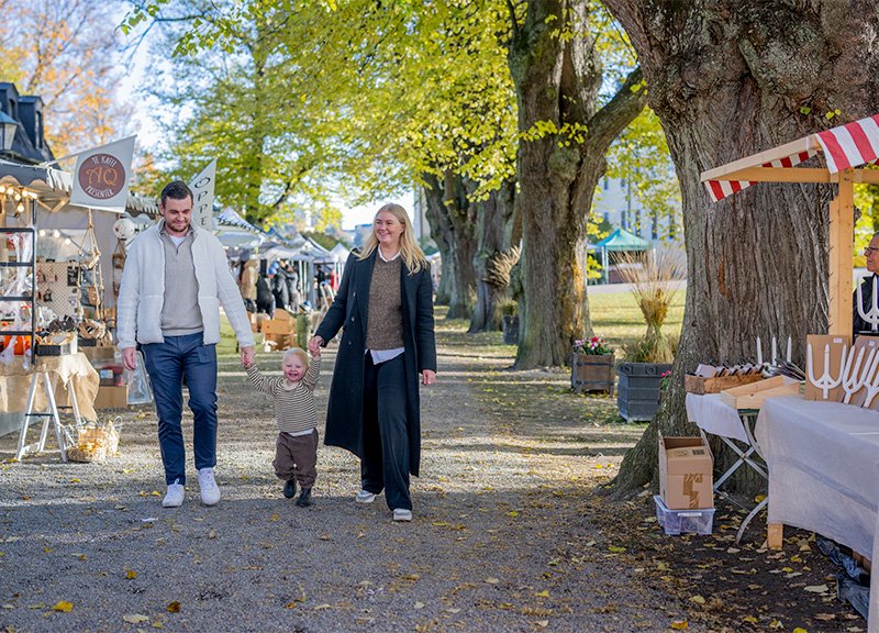 En man och en kvinna promenerar i en park med ett litet barn som håller deras händer. De passerar marknader och träd med fallfärgade löv.