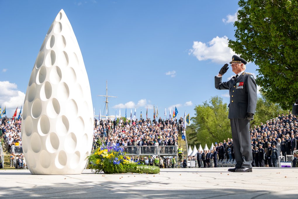 HM Karl XVI Gustaf gör honnör framför ett vitt monument under en ceremoni med stor publik i bakgrunden. Vid monumentets fot ligger en krans med blågula blommor. Bilden förmedlar högtidlighet, minne och nationell ceremoni.