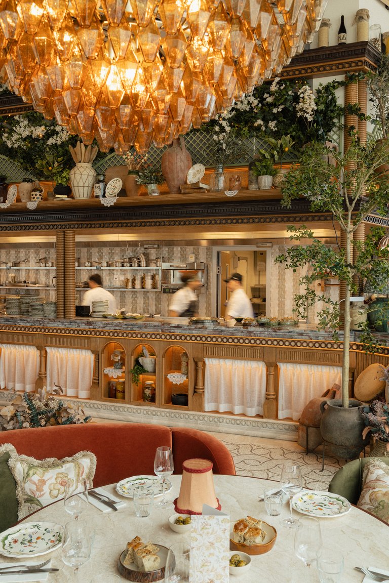 Restauranginteriör med dukat bord, öppen köksdisk, grönskande växter och en stor glaskrona i varmt ljus.