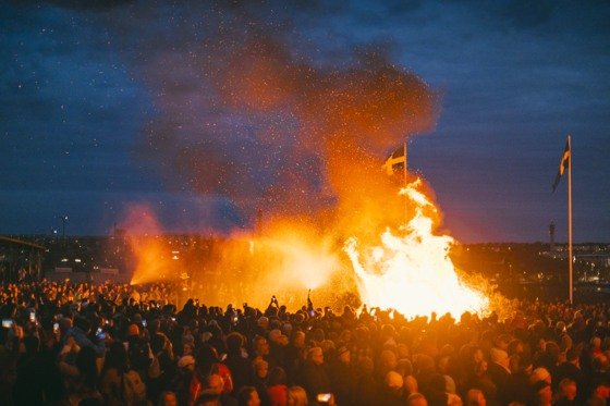 En stor folkmassa samlas runt en traditionell brasa under valborgsfirandet i Stockholm, med svenska flaggor synliga mot kvällshimlen.